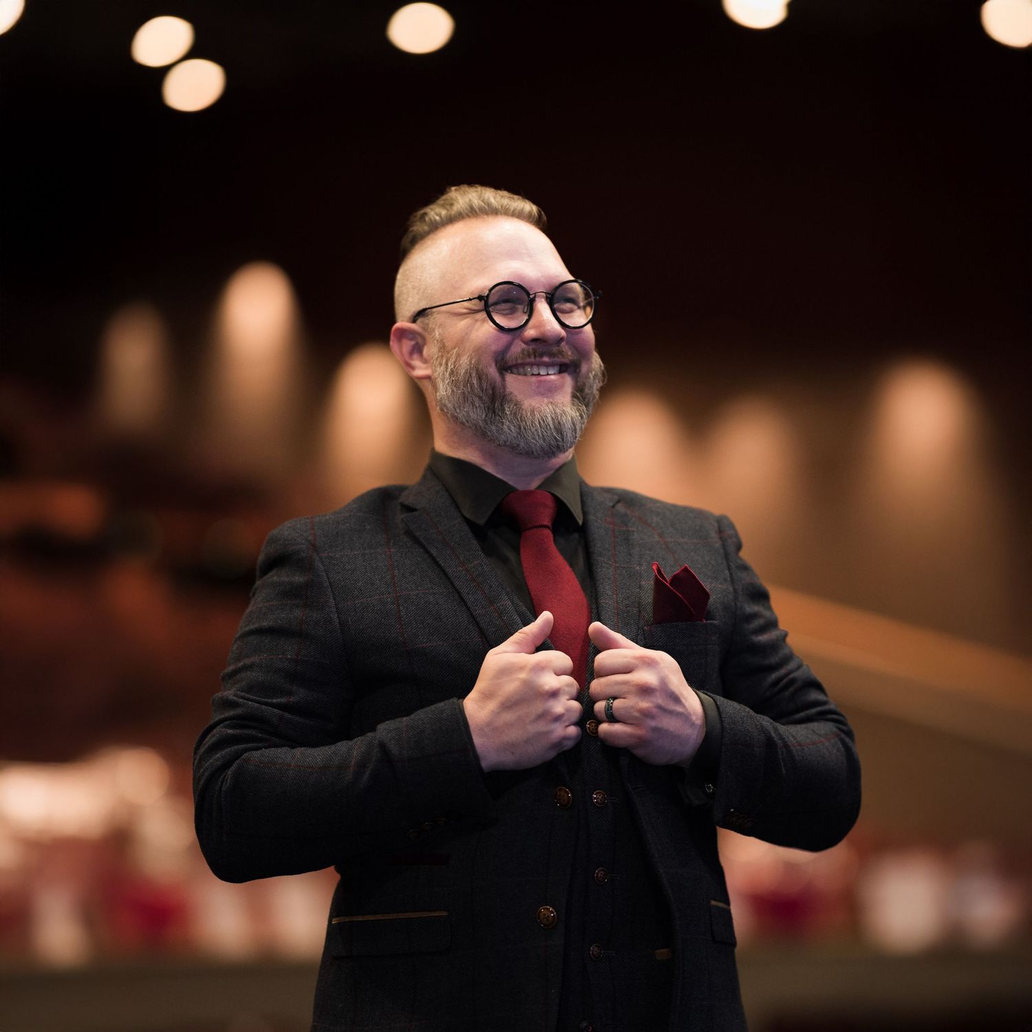 Bearded man in glasses wearing a gray and red suit, smiling in a theater with bokeh lights in the background.