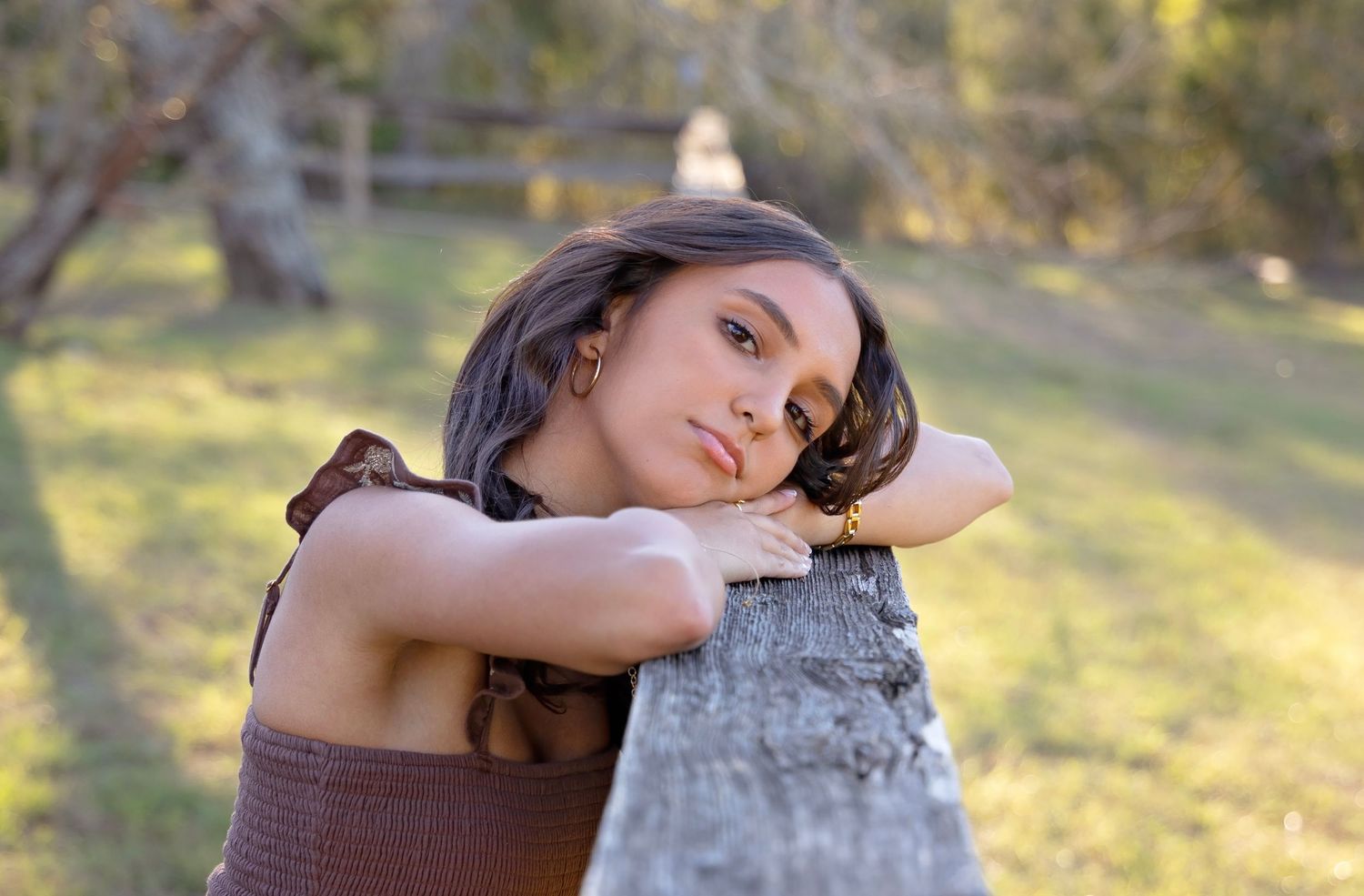 A person in brown attire poses outdoors against a tree trunk with golden sunlight filtering through the background.