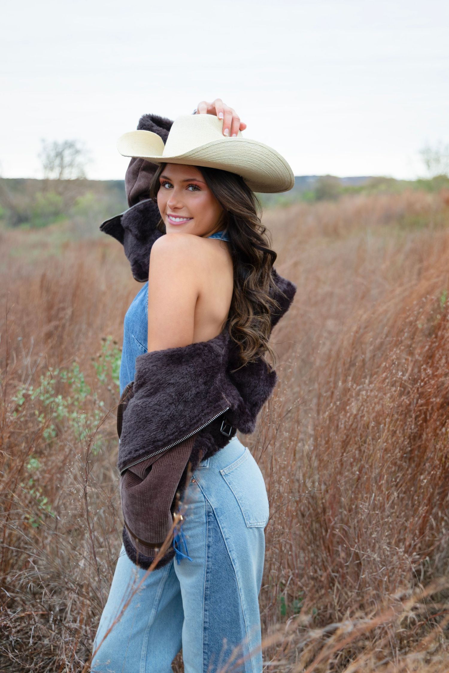 A woman in a brown hat and jeans poses playfully in a field of tall brown grass on an autumn day.