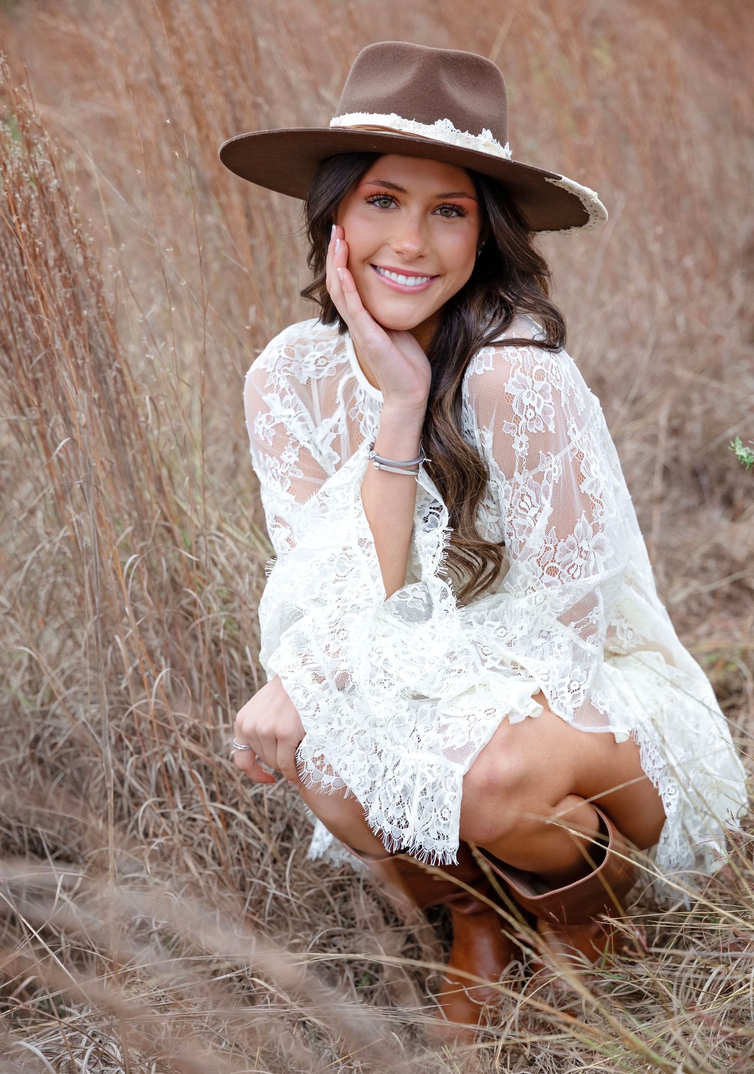 A person in a white lace dress and brown hat poses in tall brown grass during an outdoor photoshoot.