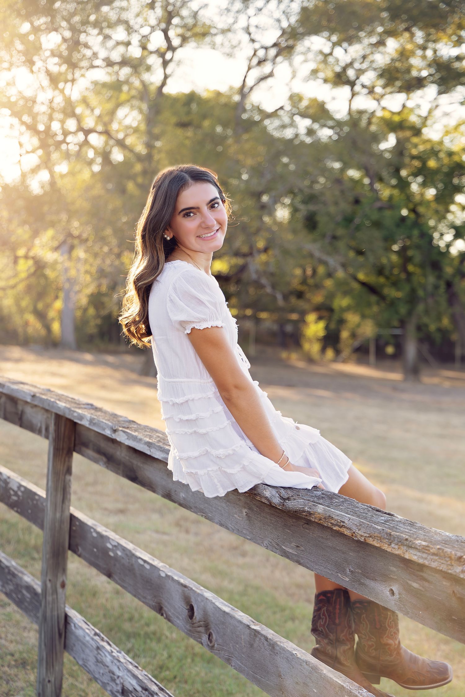 A person in a white dress sits on a rustic wooden fence at sunset in a park-like setting.