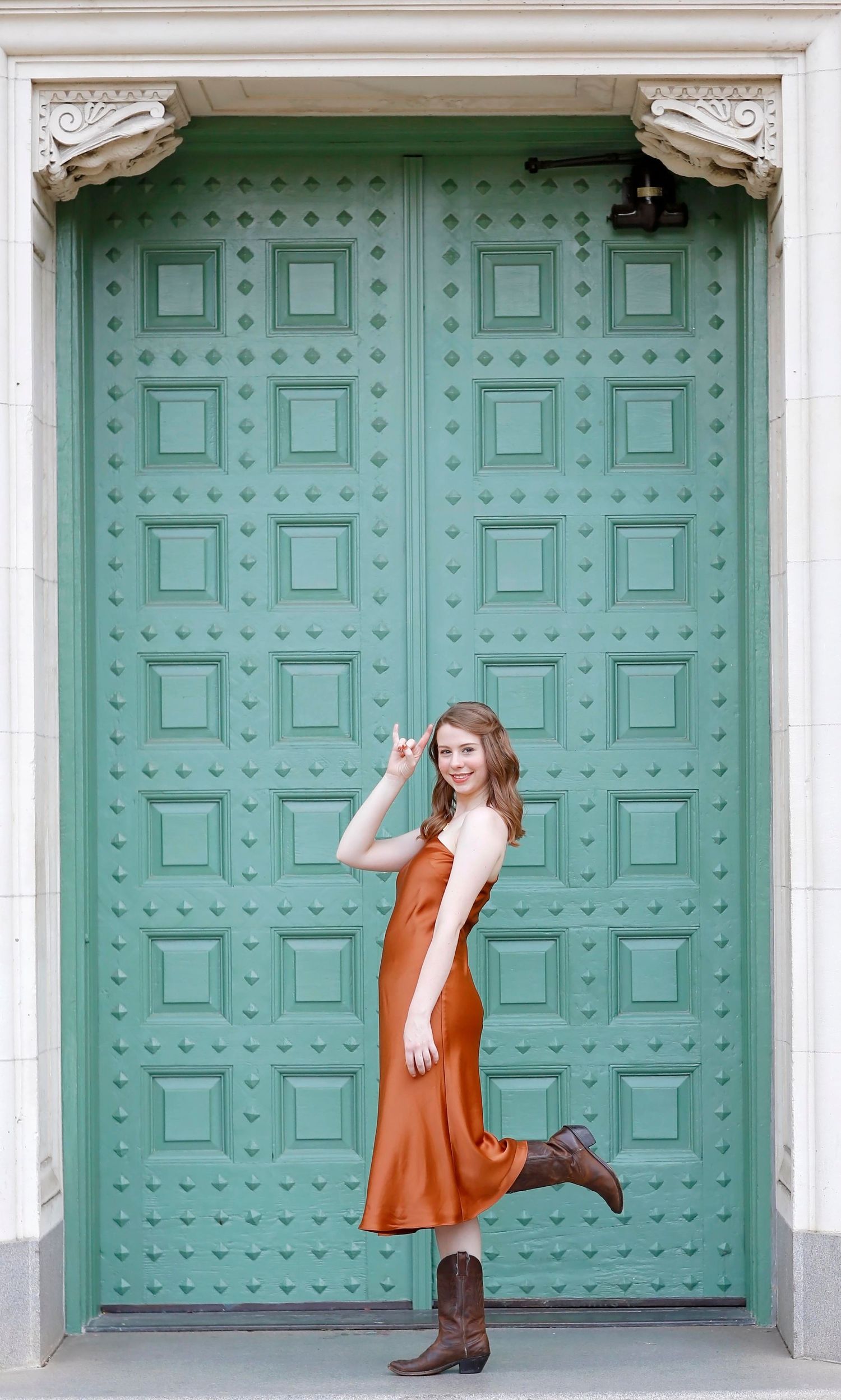 A person in a brown silk dress poses playfully against a large mint green ornate door.