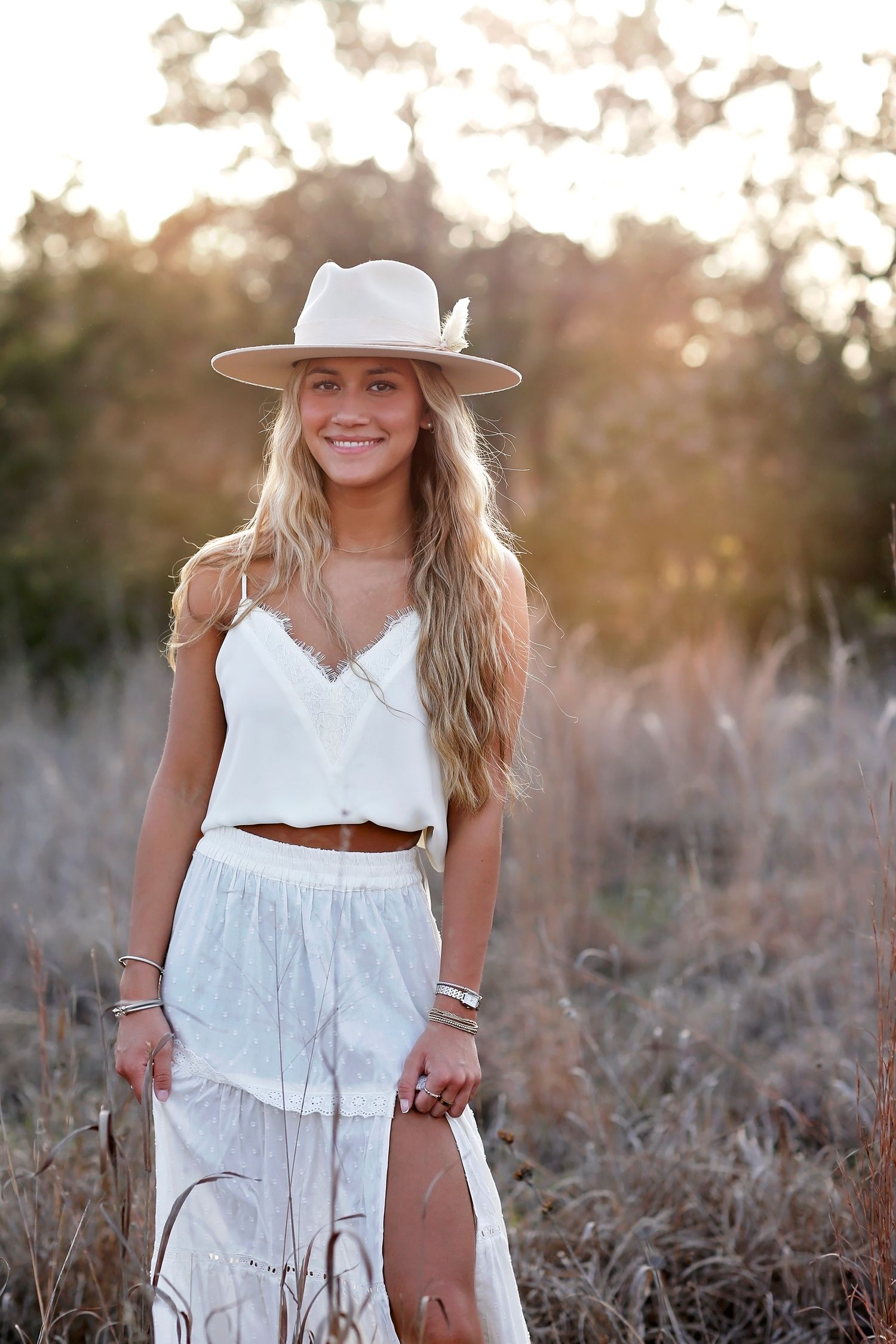 Person in white boho dress and wide brim hat poses in field during golden hour.