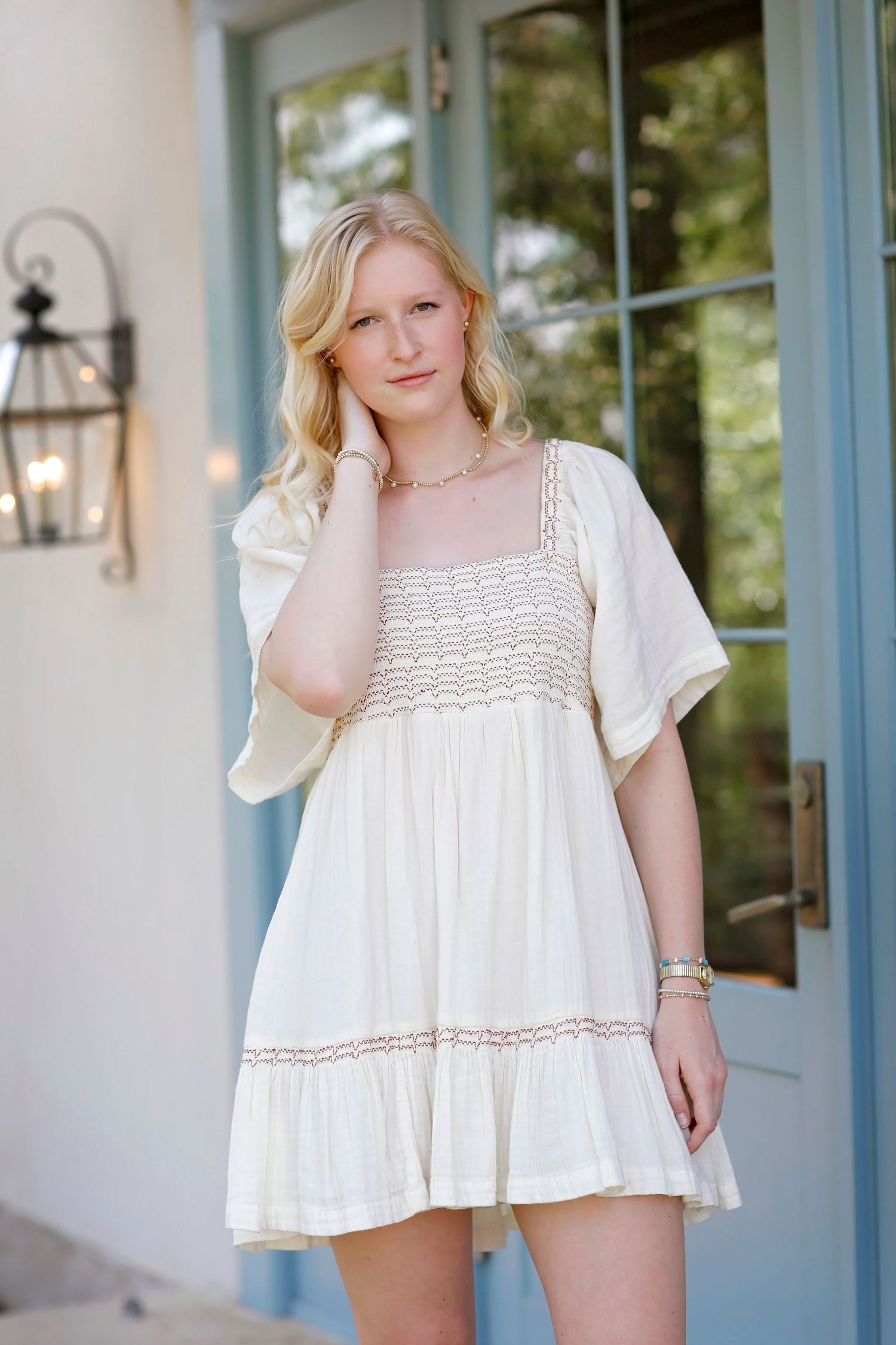 A white cotton dress with crochet lace detailing and short flutter sleeves shown in three similar poses against a blue wall.