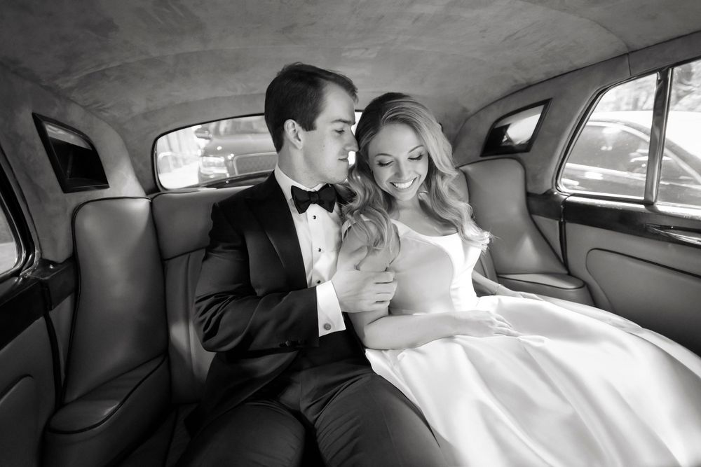 Black and white photo of a smiling bride and groom in formal attire sitting together in the backseat of a vintage car.
