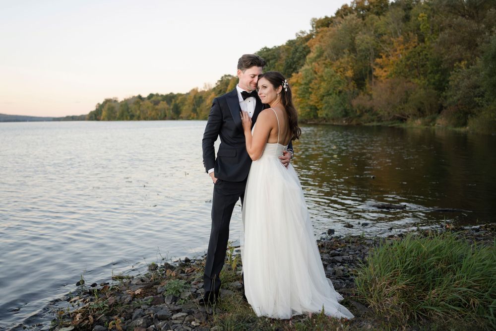 A couple in elegant wedding attire stands together by a serene lake at sunset with autumn trees in the background.
