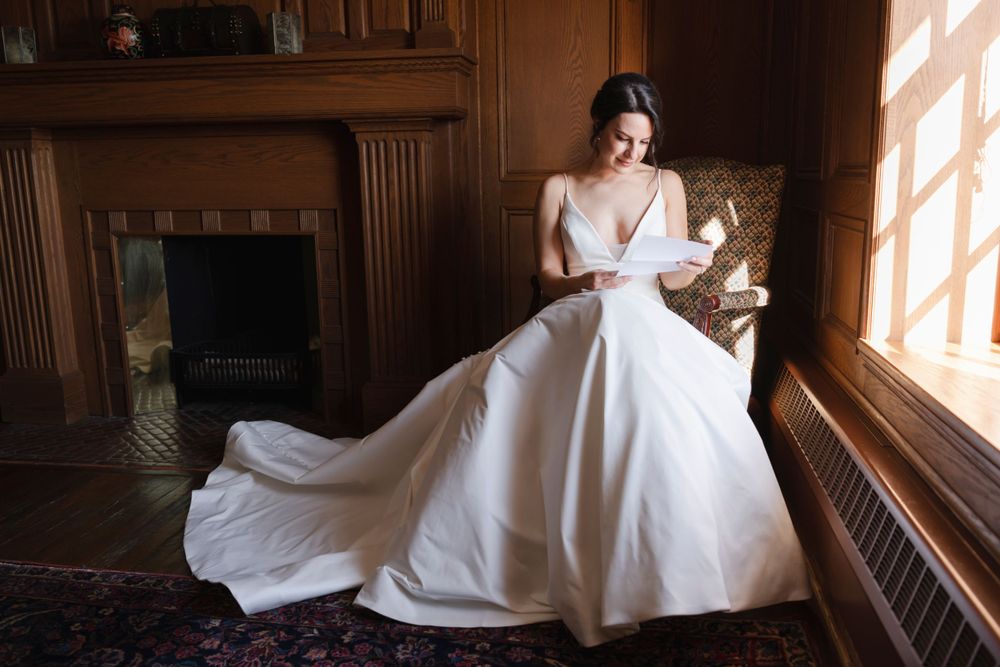 A bride in an elegant white ball gown reads a letter while seated by a window in a wood-paneled room before her wedding in Philadelphia