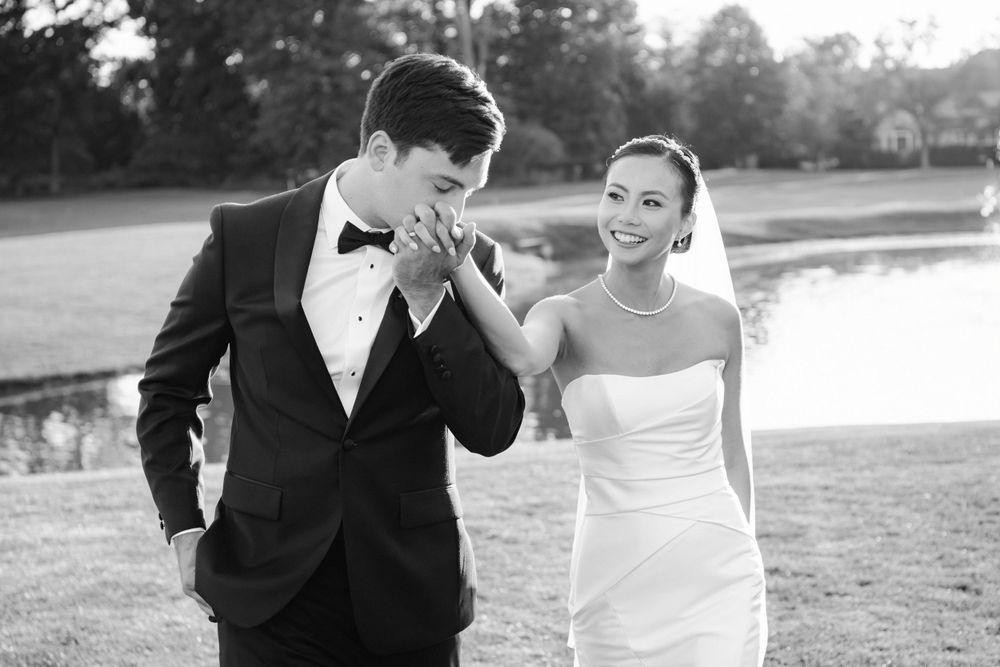 Black and white wedding photo of a couple walking together by a lake with the groom kissing the bride's hand.