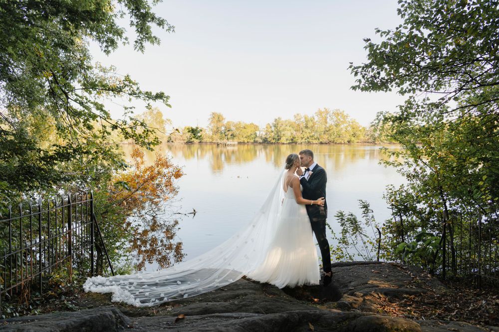 A romantic wedding couple embraces on the river's edge during golden hour, with a long flowing veil at Bartram's Garden in Philadelphia