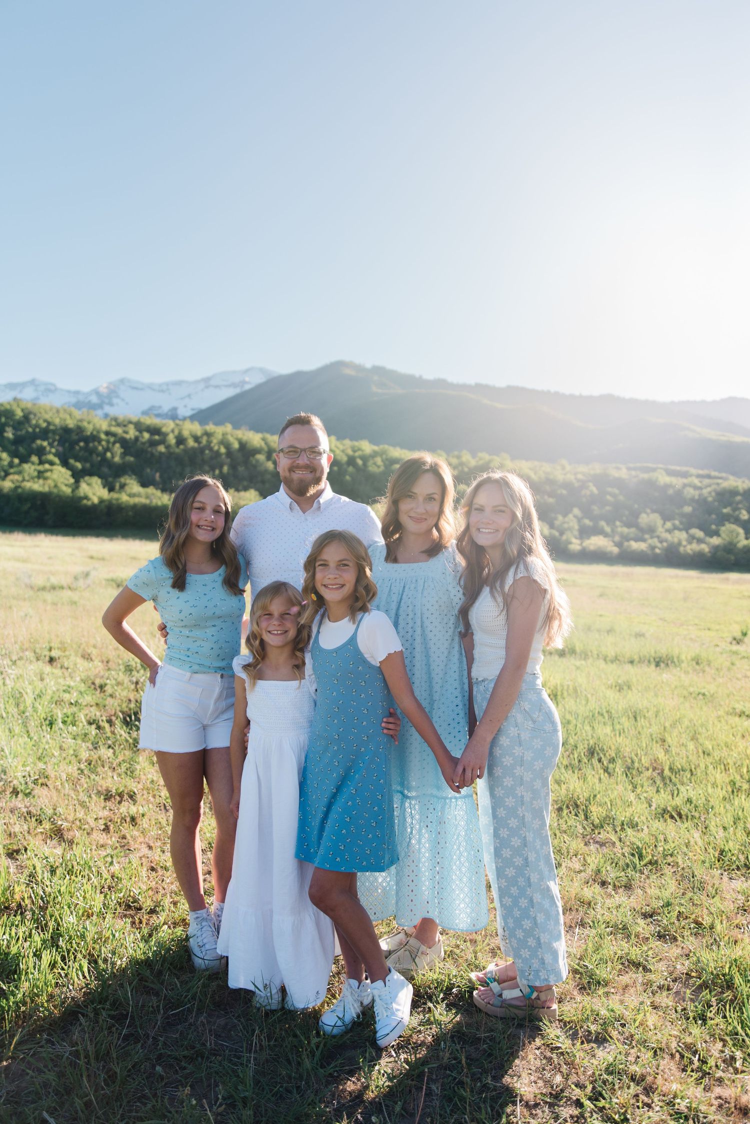 A family in coordinating blue and white outfits poses together in a sunny mountain meadow at sunset.