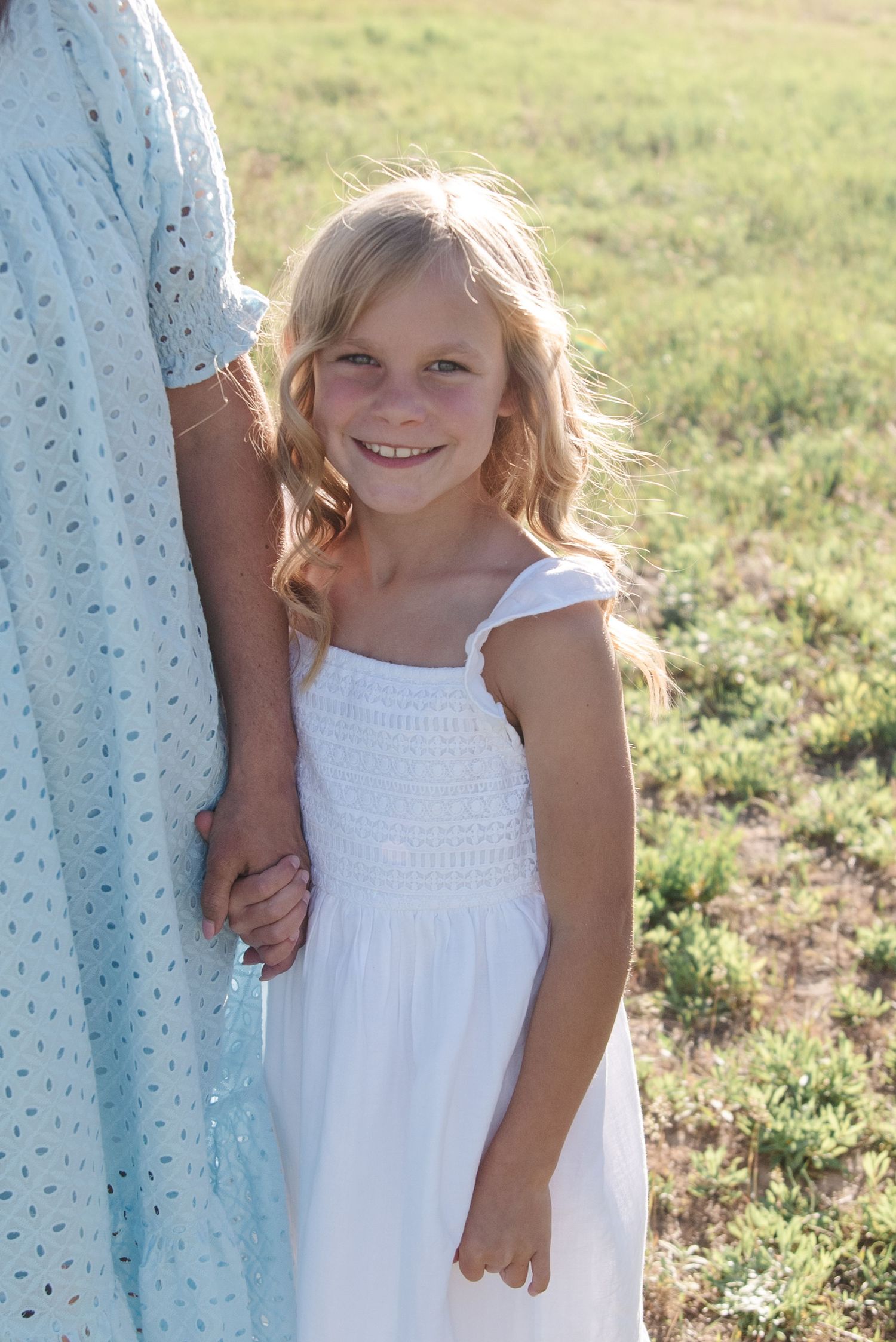 A close-up portrait of a young girl in a white dress standing outdoors in natural sunlight.
