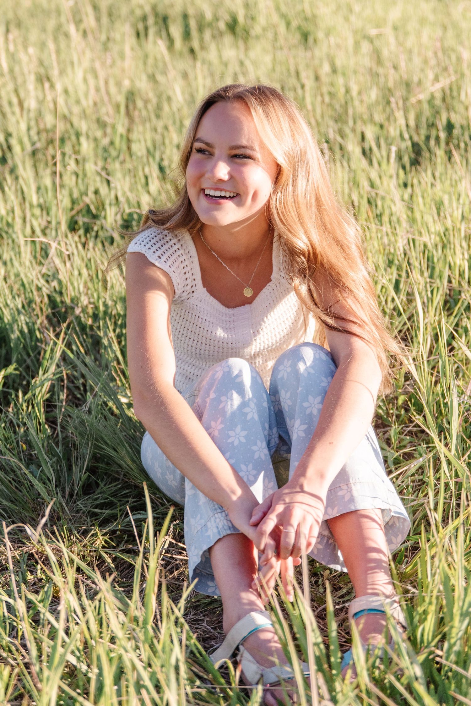 A candid portrait of someone sitting in tall grass wearing a white top and jeans, smiling brightly in sunlight.
