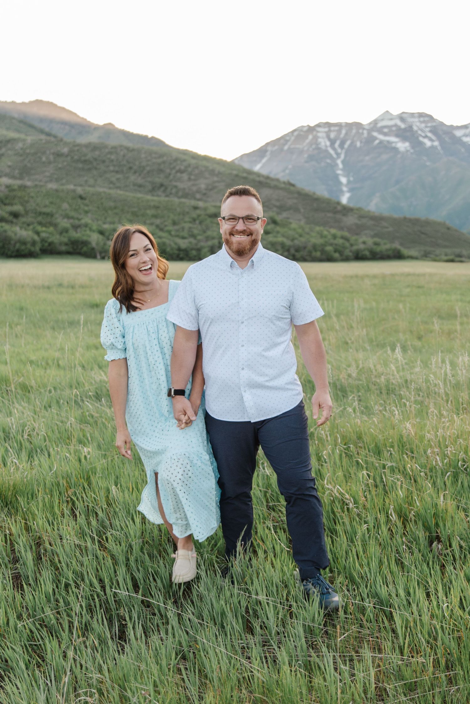A couple stands together laughing in a mountain meadow with snow-capped peaks in the background.