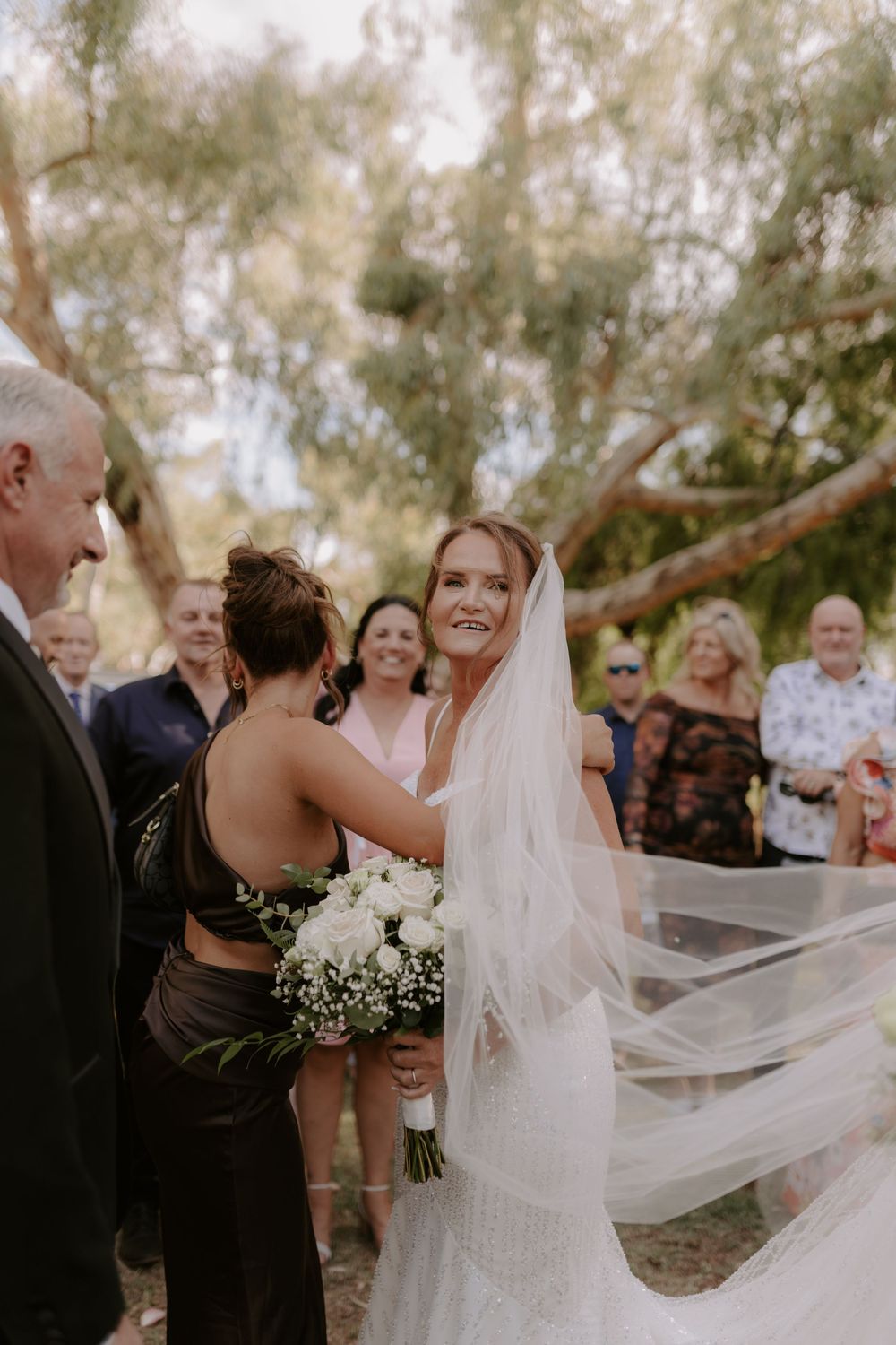 Joyful celebration with flowing white wedding dress and veil catching the sunlight in a wooded outdoor setting.