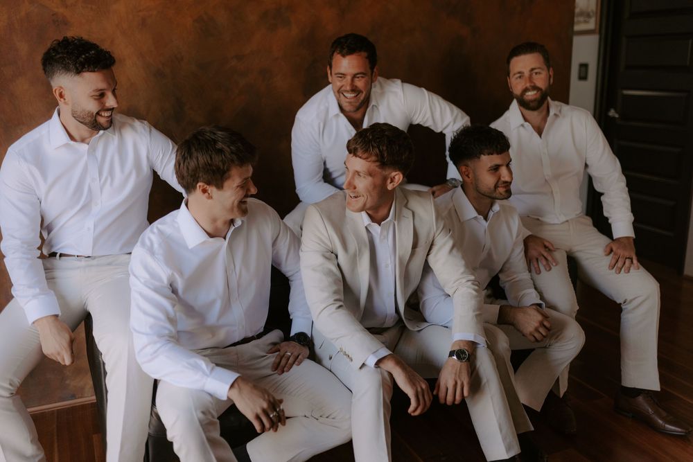 A group of groomsmen in white shirts sit together laughing and smiling during a wedding photoshoot.