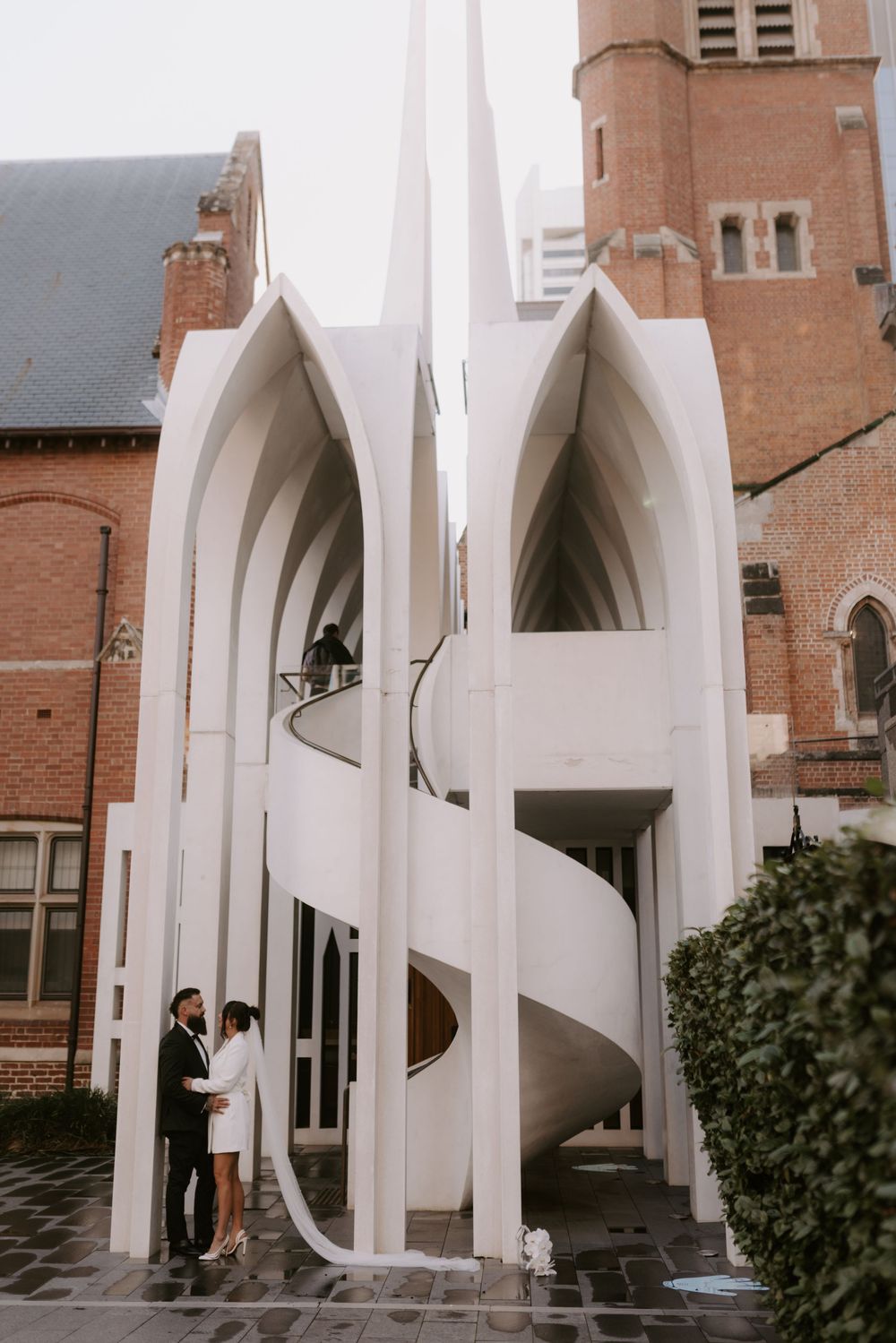 Modern white architectural arches frame a couple against a brick church backdrop.