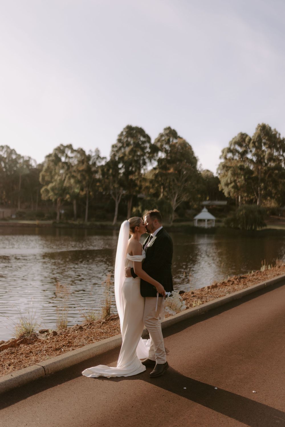 A romantic lakeside wedding photoshoot with a couple embracing on a path at sunset.