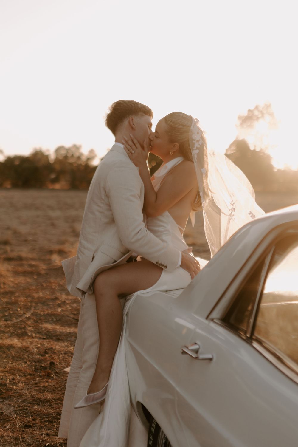 A romantic sunset moment captured between a couple in white attire next to a vintage car.