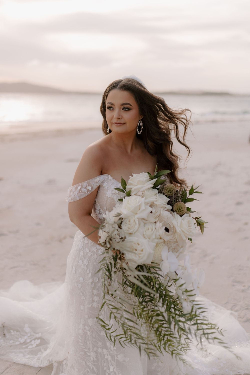 A bride in a white off-shoulder gown holds a lush bouquet of white flowers and greenery on a misty beach at sunset.