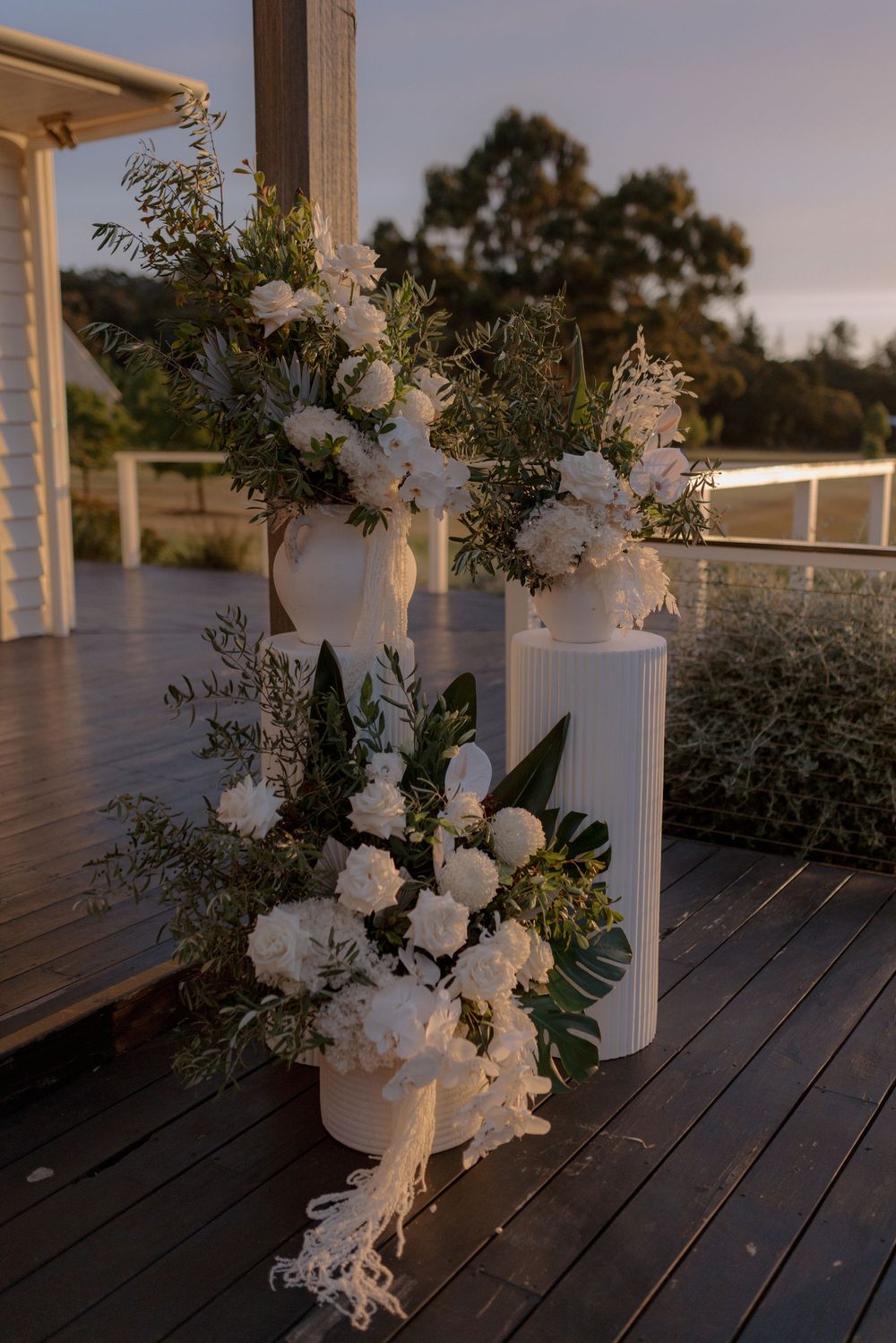 White floral arrangements in ceramic vases line a rustic wooden deck railing at sunset creating an elegant outdoor display.