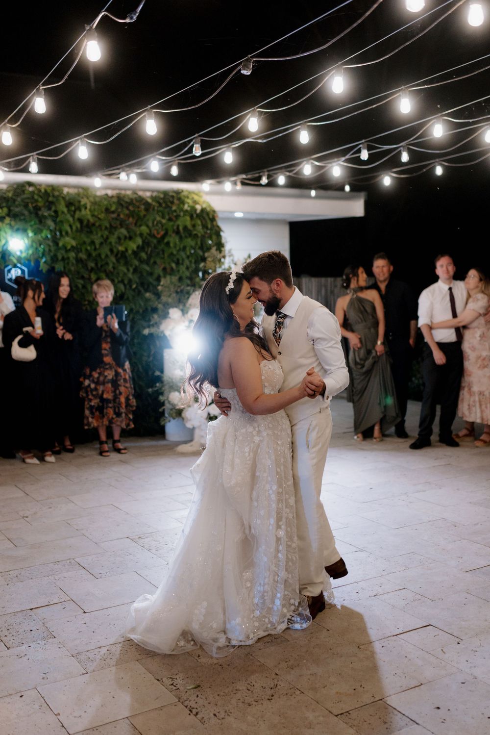 Couple sharing their first dance under string lights at an evening wedding reception with guests watching.