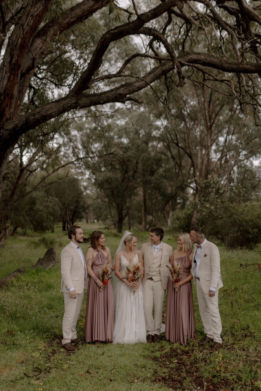 Wedding party in mauve and cream colors posing together under trees in a natural outdoor setting.
