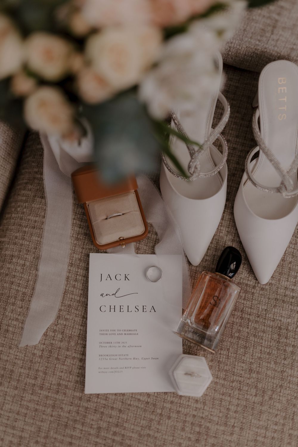 Wedding accessories including white heels, invitation cards, and perfume bottles arranged on a textured gray fabric.