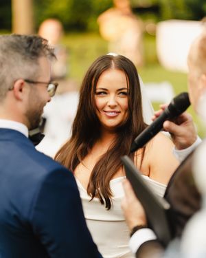 Couple exchanges vows during outdoor wedding ceremony as officiant holds microphone.