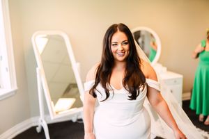 Bride wearing a white wedding dress smiles while standing in a bridal boutique with green dresses in background.