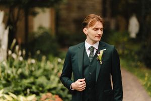 A groom in a dark suit with a yellow boutonniere stands outdoors during a wedding ceremony.