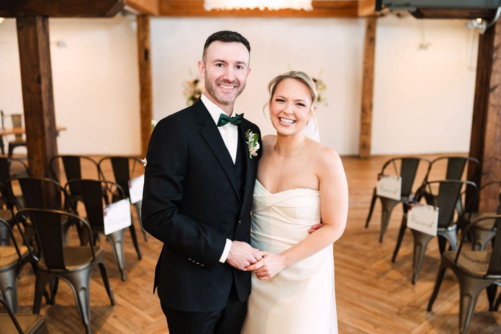 A couple in wedding attire laughs together in a rustic indoor venue with wooden floors and chairs.