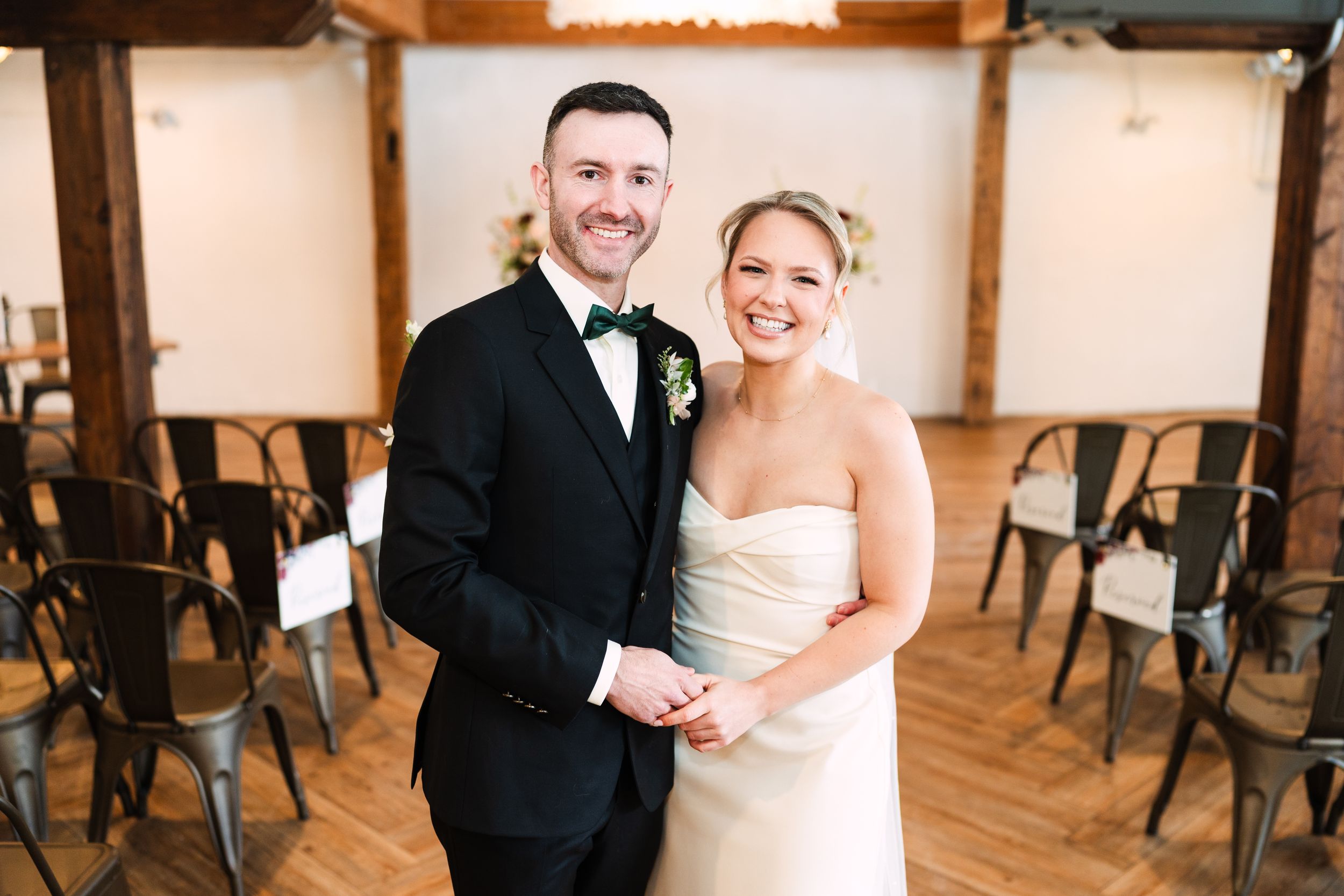 A couple in wedding attire laughs together in a rustic indoor venue with wooden floors and chairs.