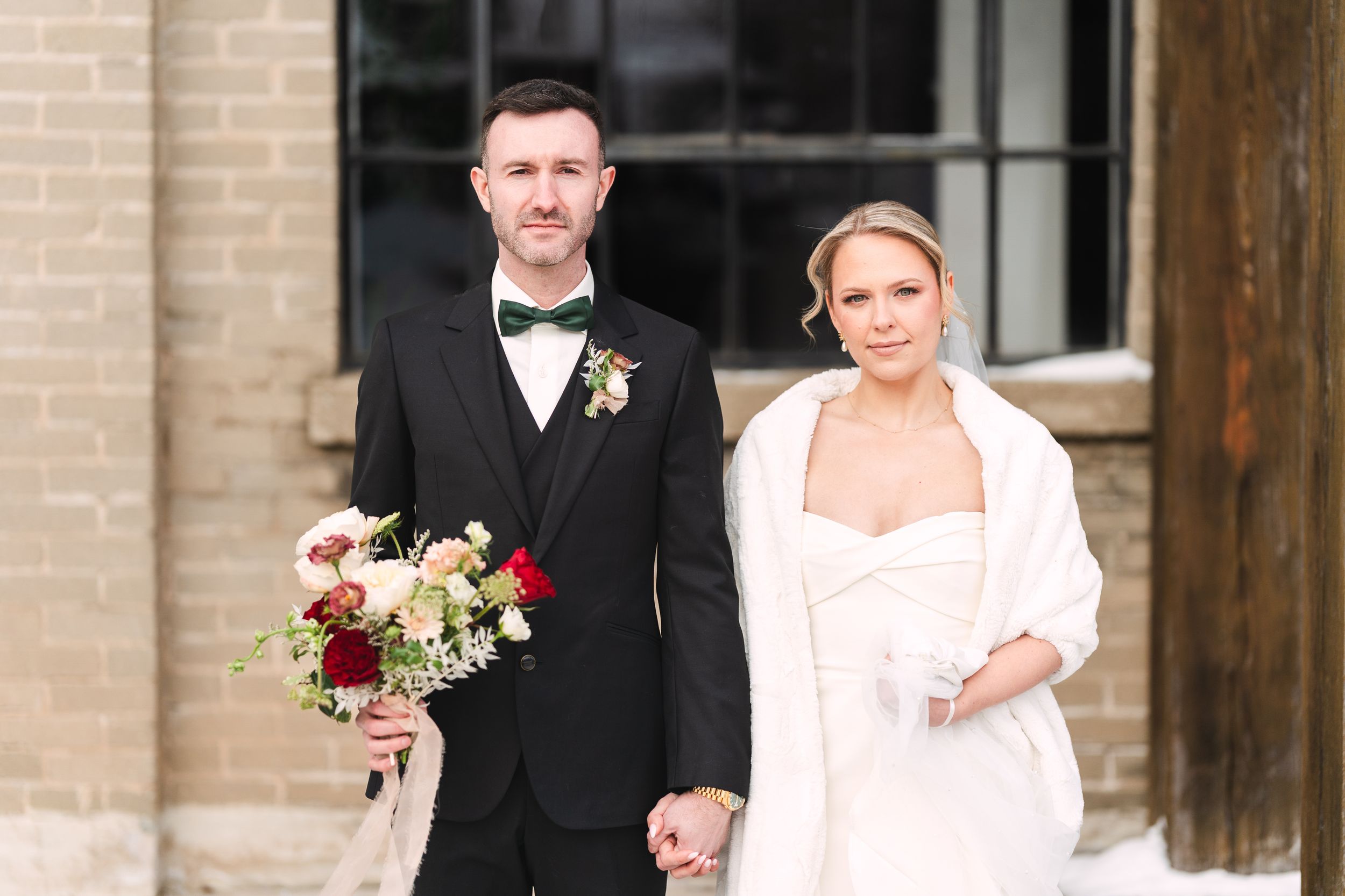 Wedding party poses together outside brick building with bride in white dress holding red and white floral bouquet.