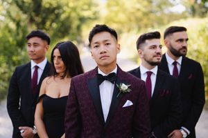 Wedding party in burgundy and black attire poses outdoors for formal photos.