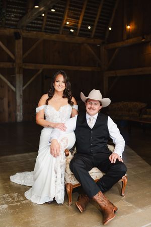 A couple in wedding attire sits together in a rustic barn setting with wooden beams and warm lighting.