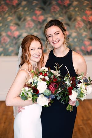 Two people in wedding attire share joyful moments while holding beautiful burgundy and white floral bouquets.