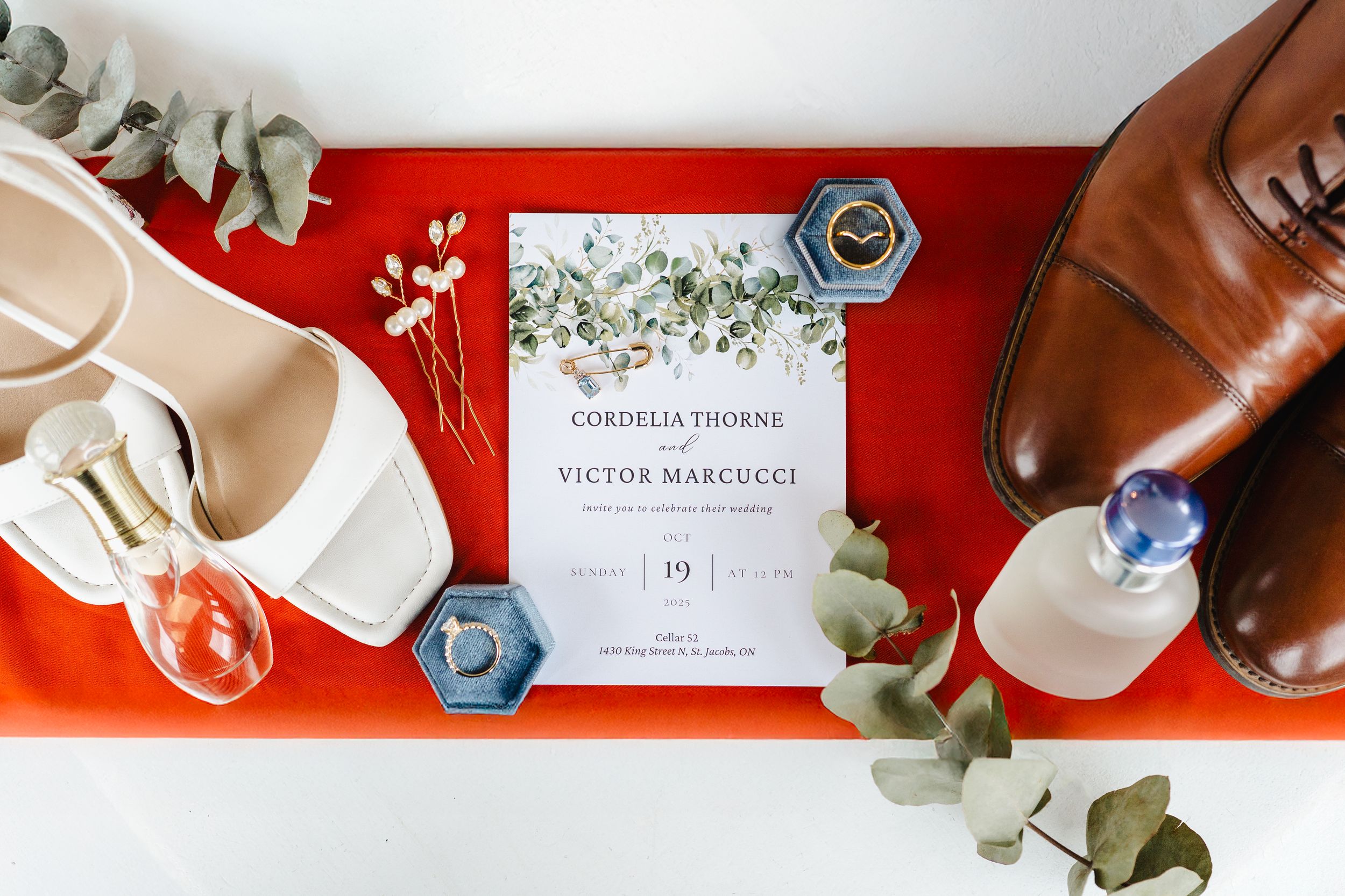 Wedding details flatlay with eucalyptus leaves, invitation cards, and brown leather shoes on red background.
