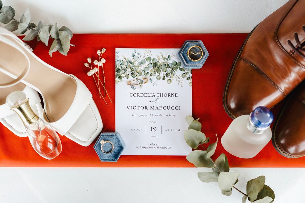 Wedding details flatlay with eucalyptus leaves, invitation cards, and brown leather shoes on red background.