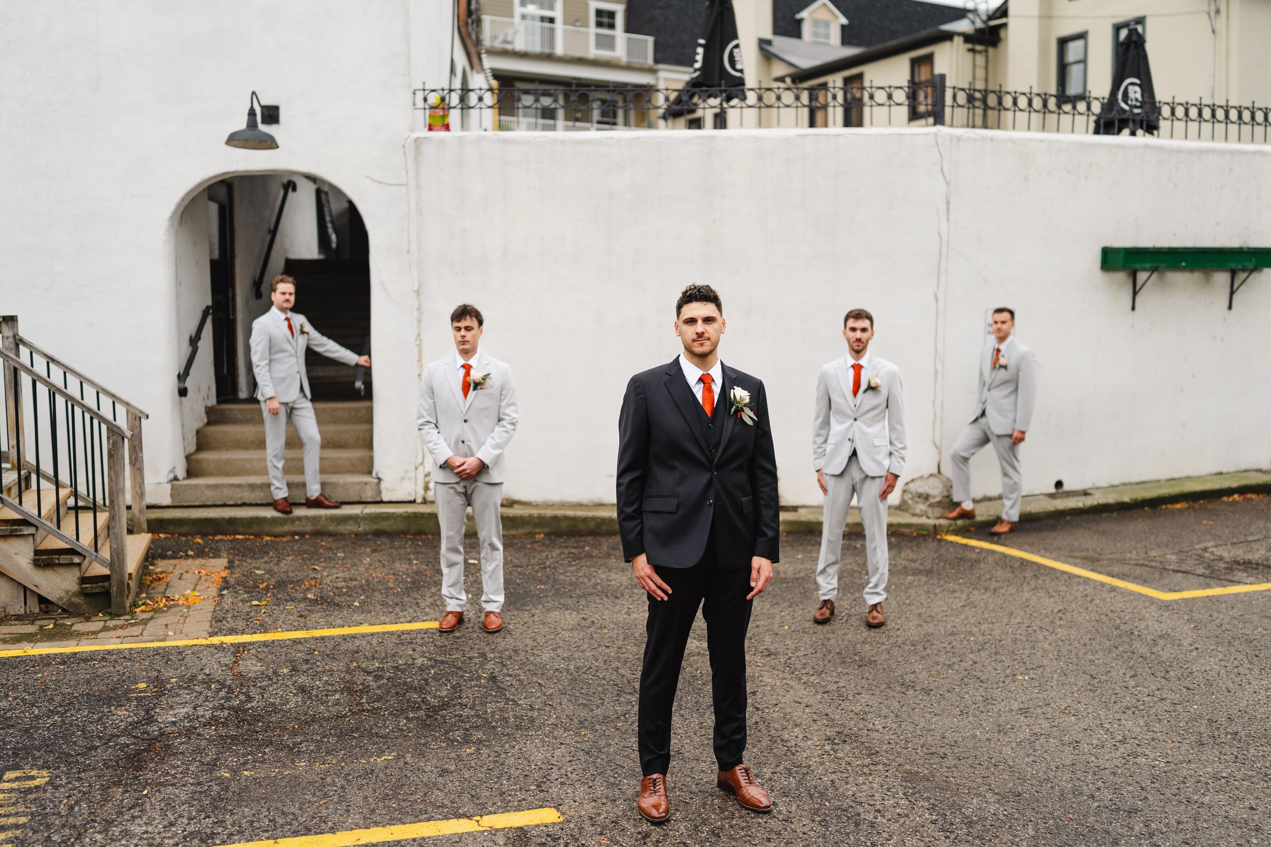 A wedding party poses together outside a building, with groomsmen in light suits and dark formal attire.