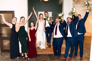 Wedding party in navy blue and burgundy attire jumps joyfully for group photos in a bright indoor venue.