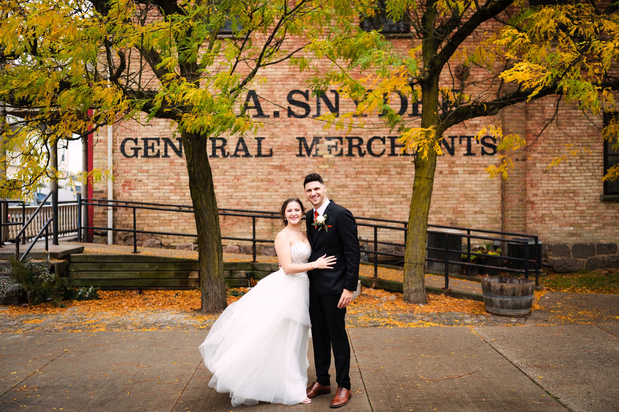 A couple walks along a rustic brick path surrounded by autumn foliage and historic buildings.