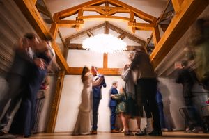 Wedding guests dance and celebrate under rustic wooden beams at an indoor reception venue.