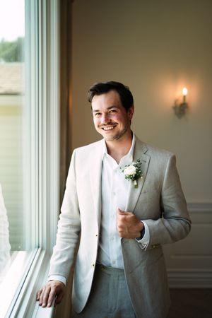 A groom in a light gray suit and white boutonniere stands by a window with warm lighting.