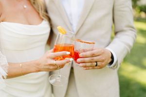 Couple holding orange cocktails during wedding celebration outdoors.