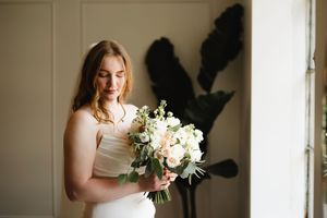 A bride in a white dress holds a delicate bouquet of white flowers while standing in soft window light.