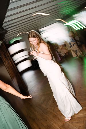 A person in a white formal dress holds a drink while dancing on a wooden floor with green lighting effects.