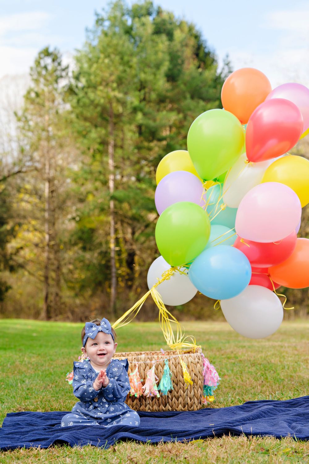 Colorful balloons tied to picnic basket on blue blanket in grassy field with trees in background for one year portrait session in Ohio