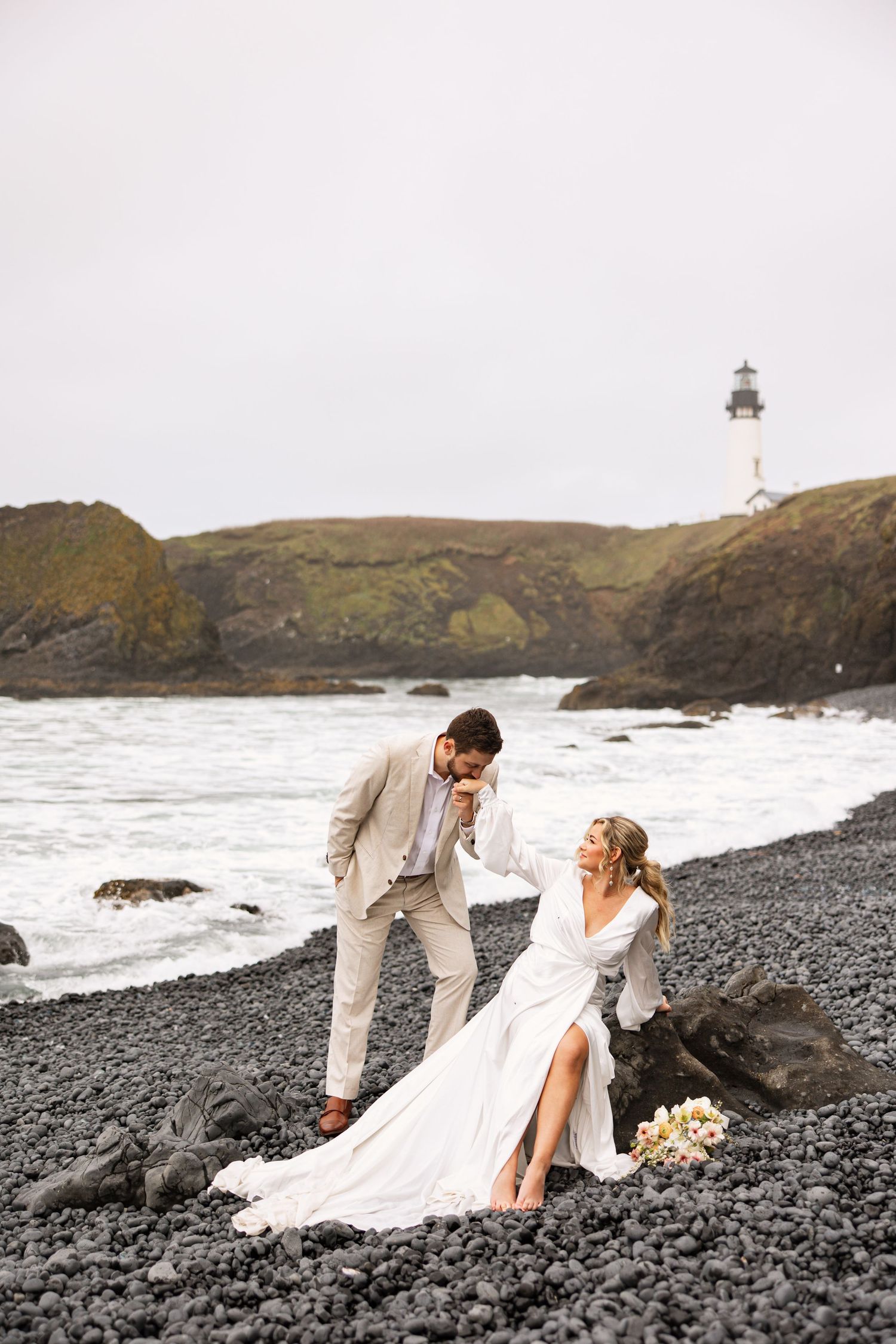 A couple in wedding attire sits on a rocky beach with a lighthouse visible on the cliff in the background.