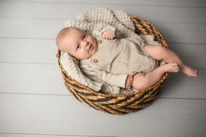 Infant lying in woven basket with cream knit blanket on white wooden floor.