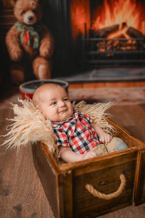 Baby wearing red plaid shirt lying in rustic wooden box with straw near fireplace.